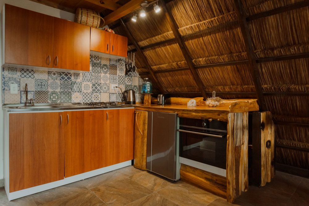 Cocina en madera con estufa, horno, lavaplatos, cafetera, nevera y buena iluminación. Chalet de Paja, Luciana Cabañas Boutique.